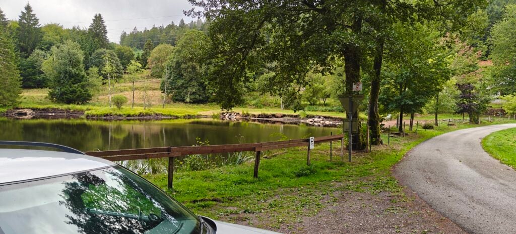 A lake with a road next to it. By the road, there's a gravel area, where a car is parked. 