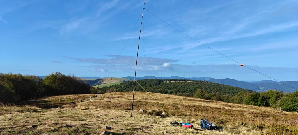 A grassy hilltop with some trees. Other hills are in the distance. My radio aerial mast and backpack are in the foreground.