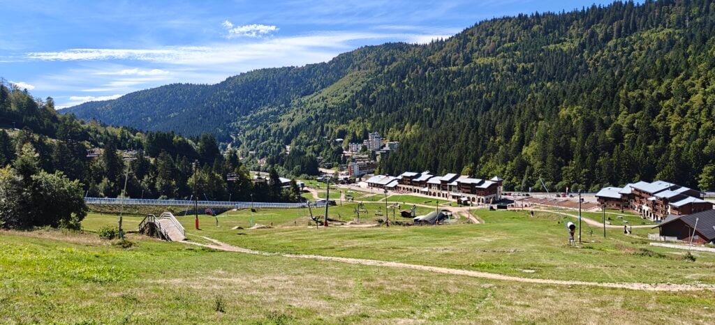 A green valley between forested hills. There are many buildings in the valley. The hill in the foreground has the apparatus of a ski resort, including chair lifts and snow blowers. 